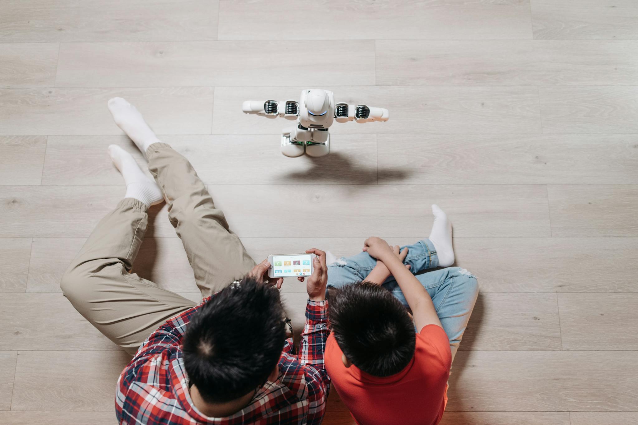 A father and son using a smartphone to control a small humanoid robot on wooden flooring.