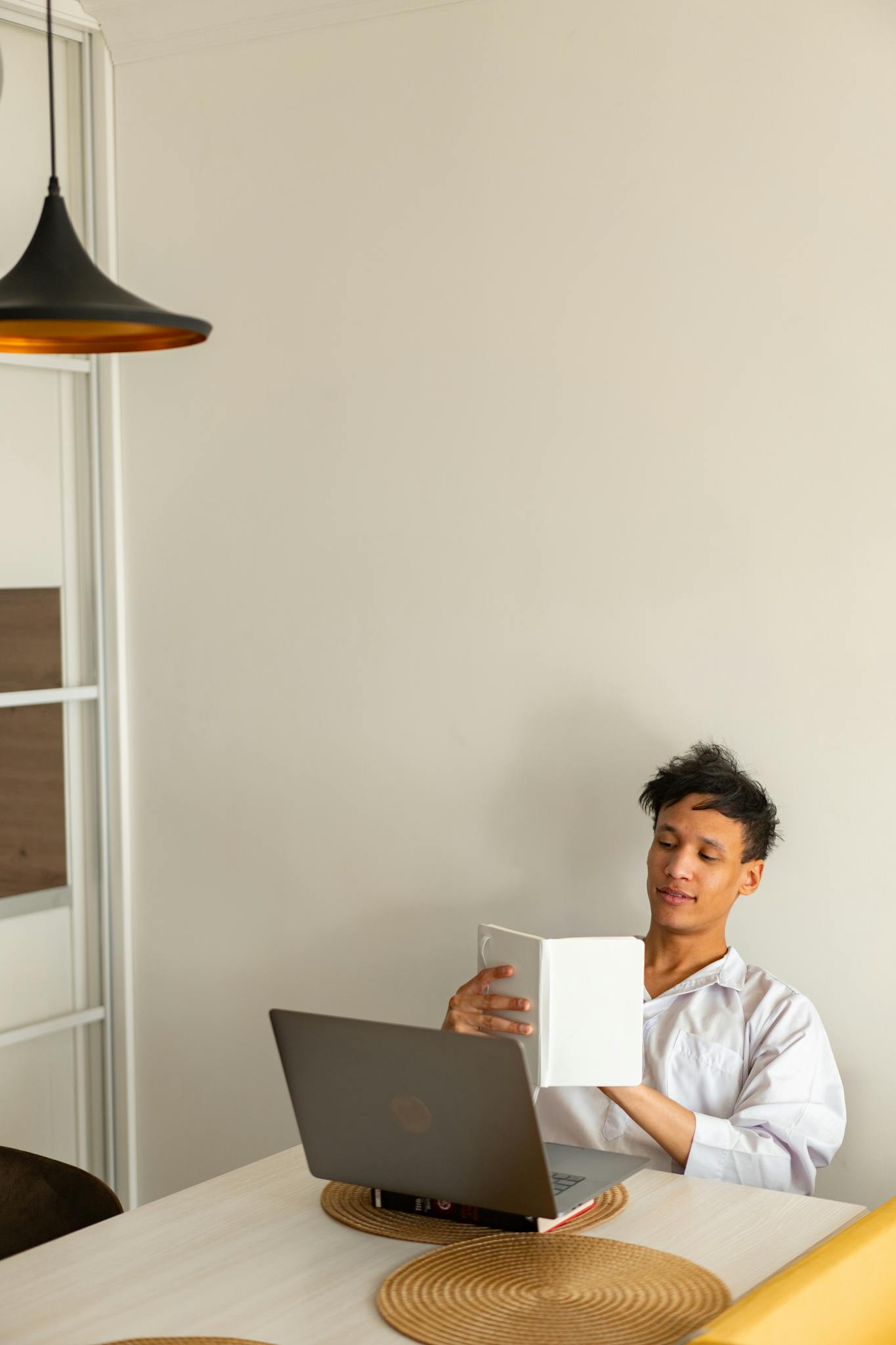 A young man studies in a minimalist home office with a laptop and book.